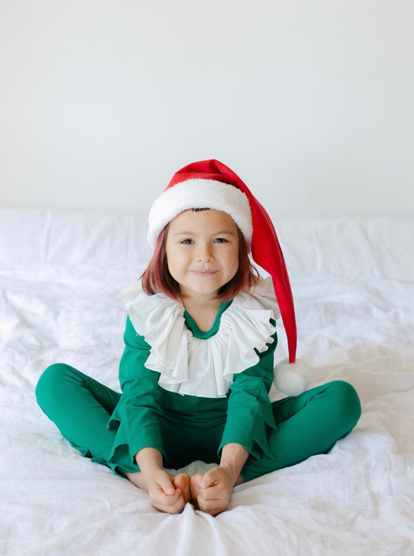 Child wearing a green Christmas elf pajama costume with frilled collar and red Santa hat, sitting on a white bed
