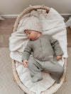 Baby in a green outfit and knit hat lying in a wicker crib with white bedding.
