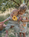 Two children outdoors, one holding a baby and the other holding a plush toy, with trees in the background.
