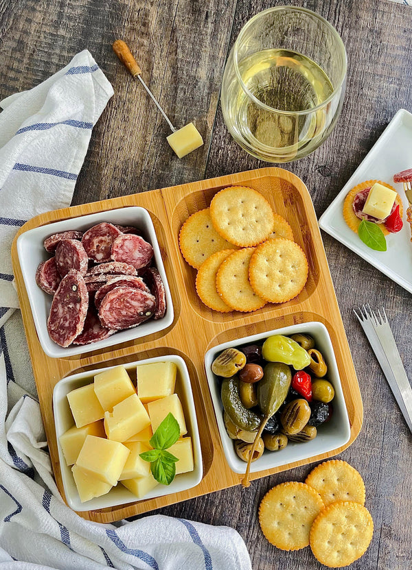 A natural bamboo square divided dish with four compartments, each filled with different food items such as crackers and cheese, displayed on a table with a glass of wine and cutlery.