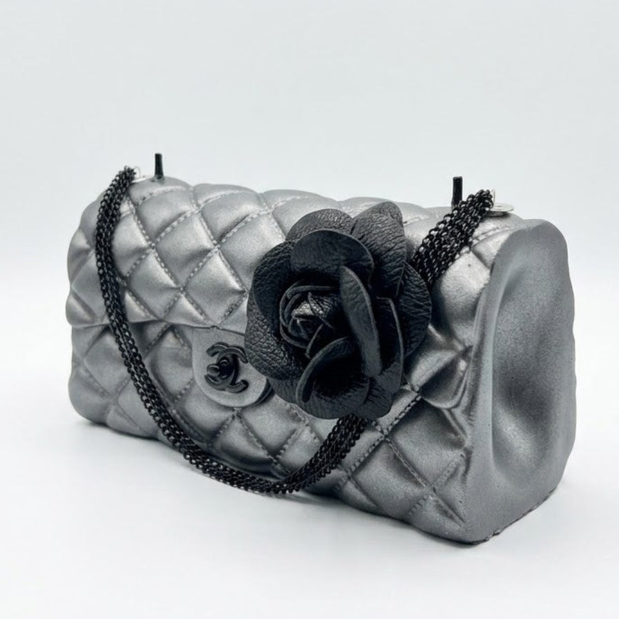 A metallic silver quilted handbag with a black chain strap and a large black fabric flower embellishment on the front, set against a plain light gray background.