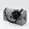 A metallic silver quilted handbag with a black chain strap and a large black fabric flower embellishment on the front, set against a plain light gray background.
