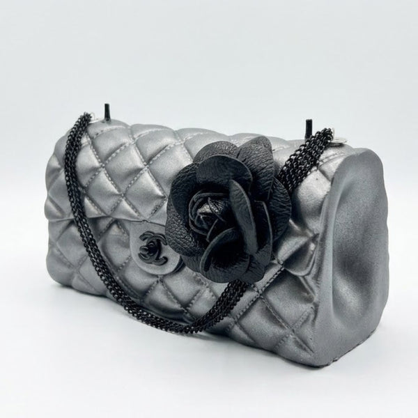 A metallic silver quilted handbag with a black chain strap and a large black fabric flower embellishment on the front, set against a plain light gray background.