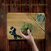 Hand holding green dough on a wooden cutting board with measuring spoons and a bowl of dried herbs.