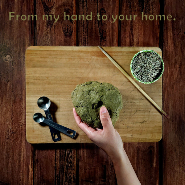 Hand holding green dough on a wooden cutting board with measuring spoons and a bowl of dried herbs.