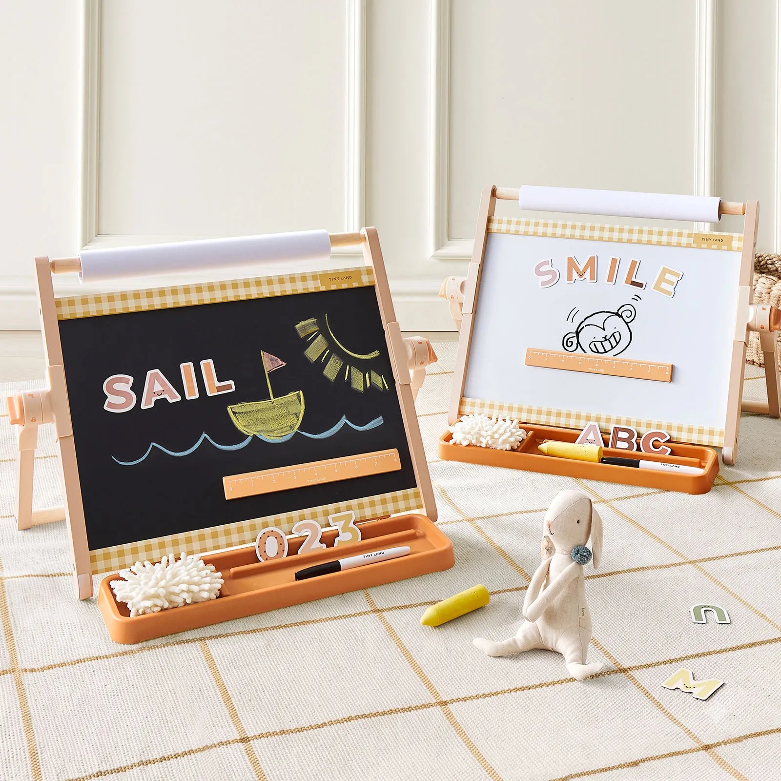 Two wooden magnetic drawing boards displayed on a beige checkered rug in a bright room. The left board is black with chalk-style drawings reading 'SAIL', and the right board is white with 'SMILE' and 'ABC'.