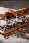 Two jars of Fine & Raw chocolate hazelnut butter on a wooden board with bread and nuts.