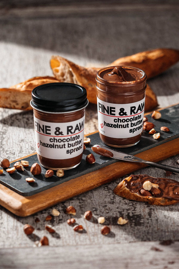 Two jars of Fine & Raw chocolate hazelnut butter on a wooden board with bread and nuts.