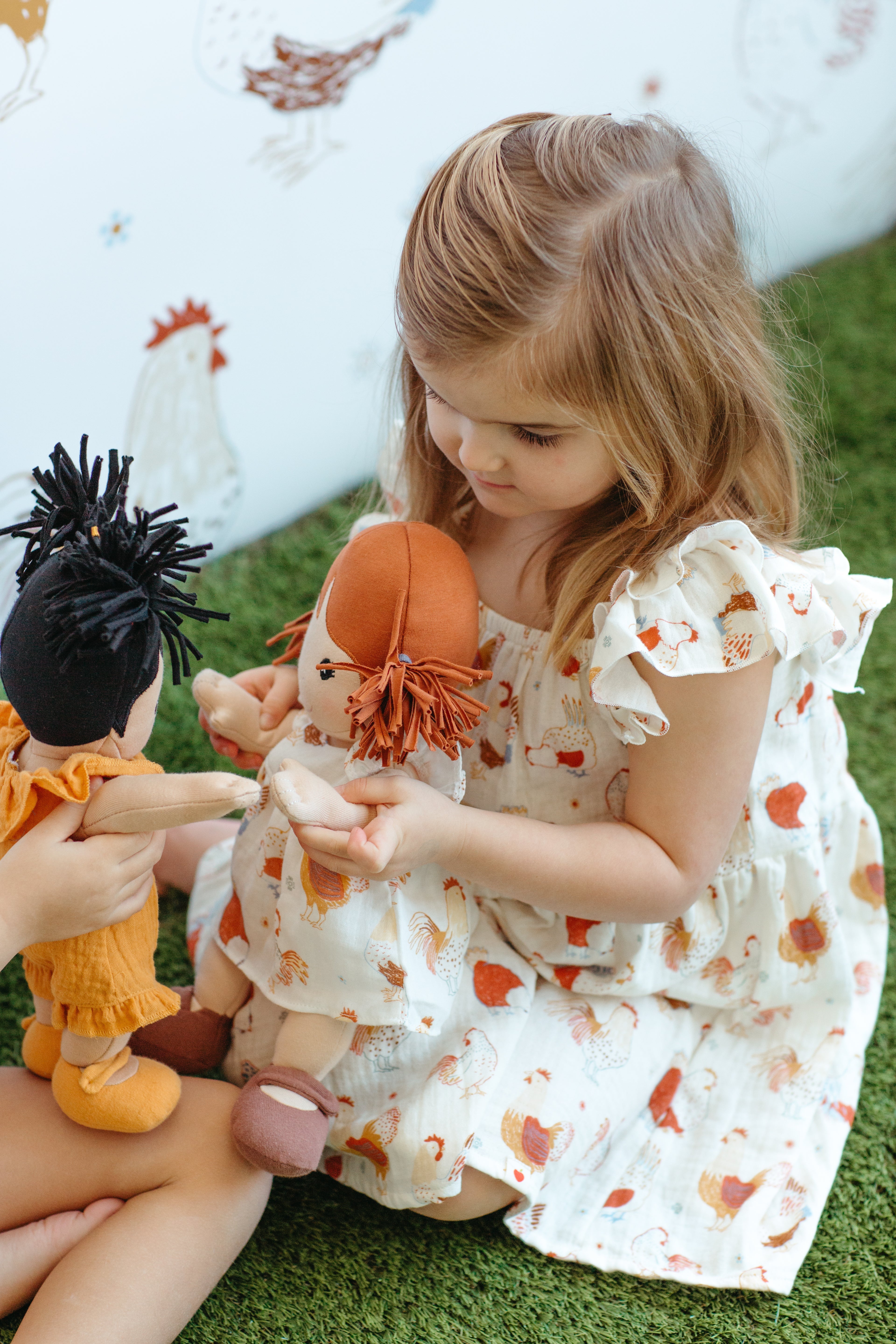 Child holding Apple Park Feathered Friends doll dress showing charming hen illustration print and flutter sleeve detail