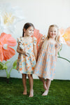 Baby wearing GOTS-certified organic muslin dress with colourful fruit print, sitting outdoors in summer