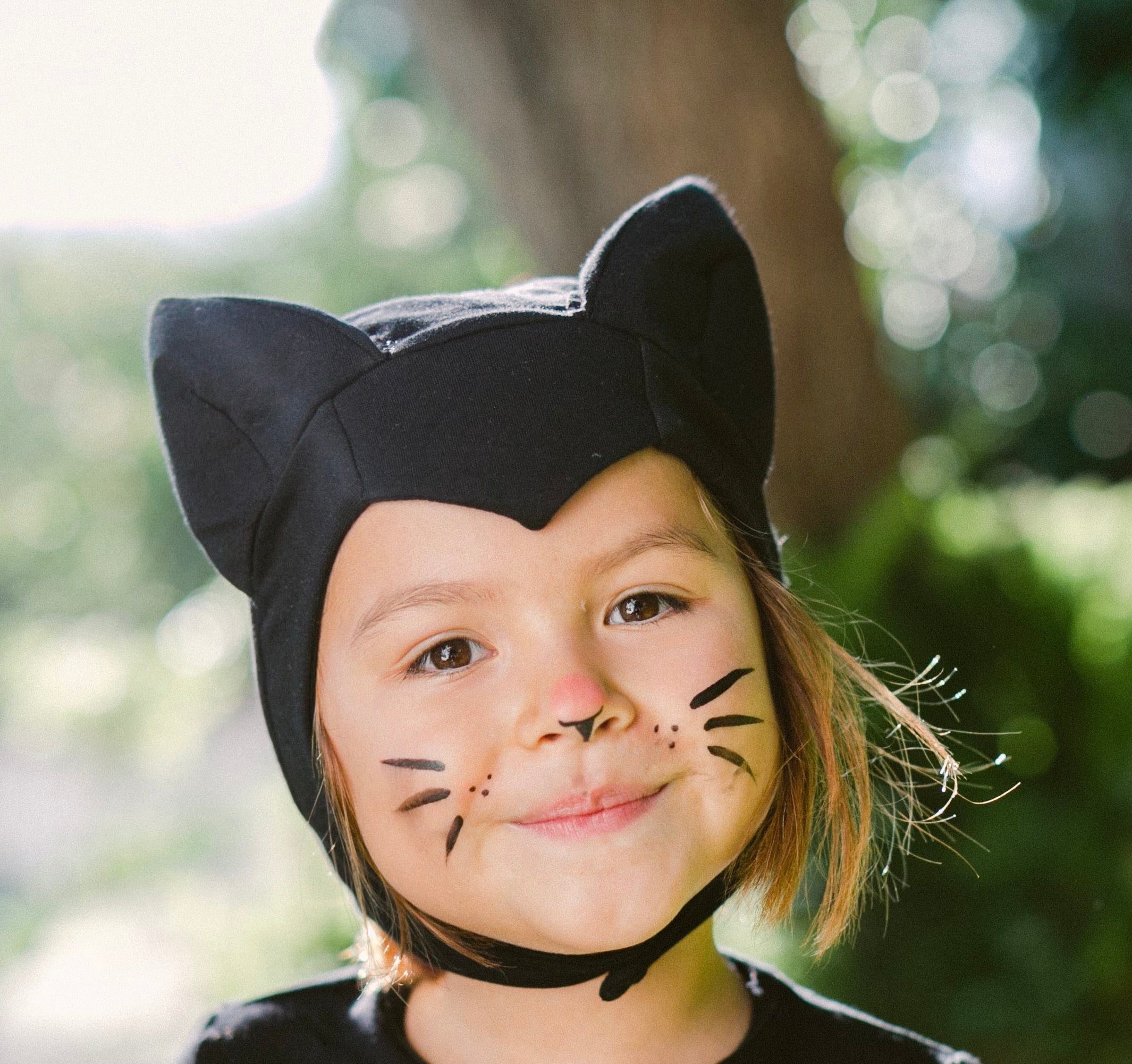 Child wearing a black cat helmet with face paint, outdoors.