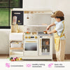 A child wearing a toy chef hat, light blue shirt, and yellow striped apron plays with a cream-colored wooden toy kitchen set. The kitchen features a sink with a gold faucet, a stove with toy food, shelves, and a small refrigerator.