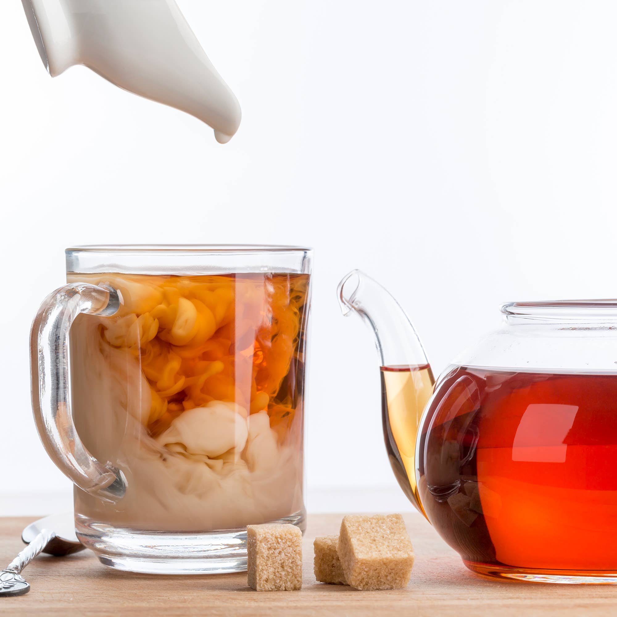 Glass mug with iced tea and lemon, teapot with hot tea, sugar cubes on a wooden surface.