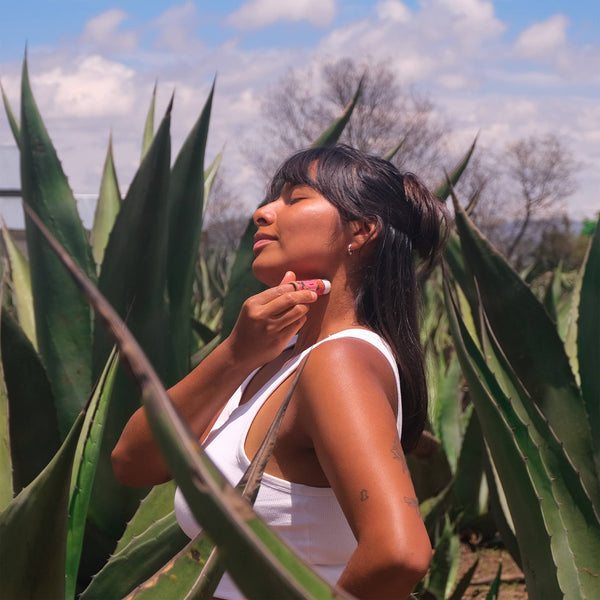 Woman applying lipstick among large green leaves with a blue sky in the background. NANAH Vanilla Coconut solid perfume — natural essential oils, lotus flower & sandalwood. Phthalate-free, vegan & longer-lasting than spray. Perfect fragrance gift.