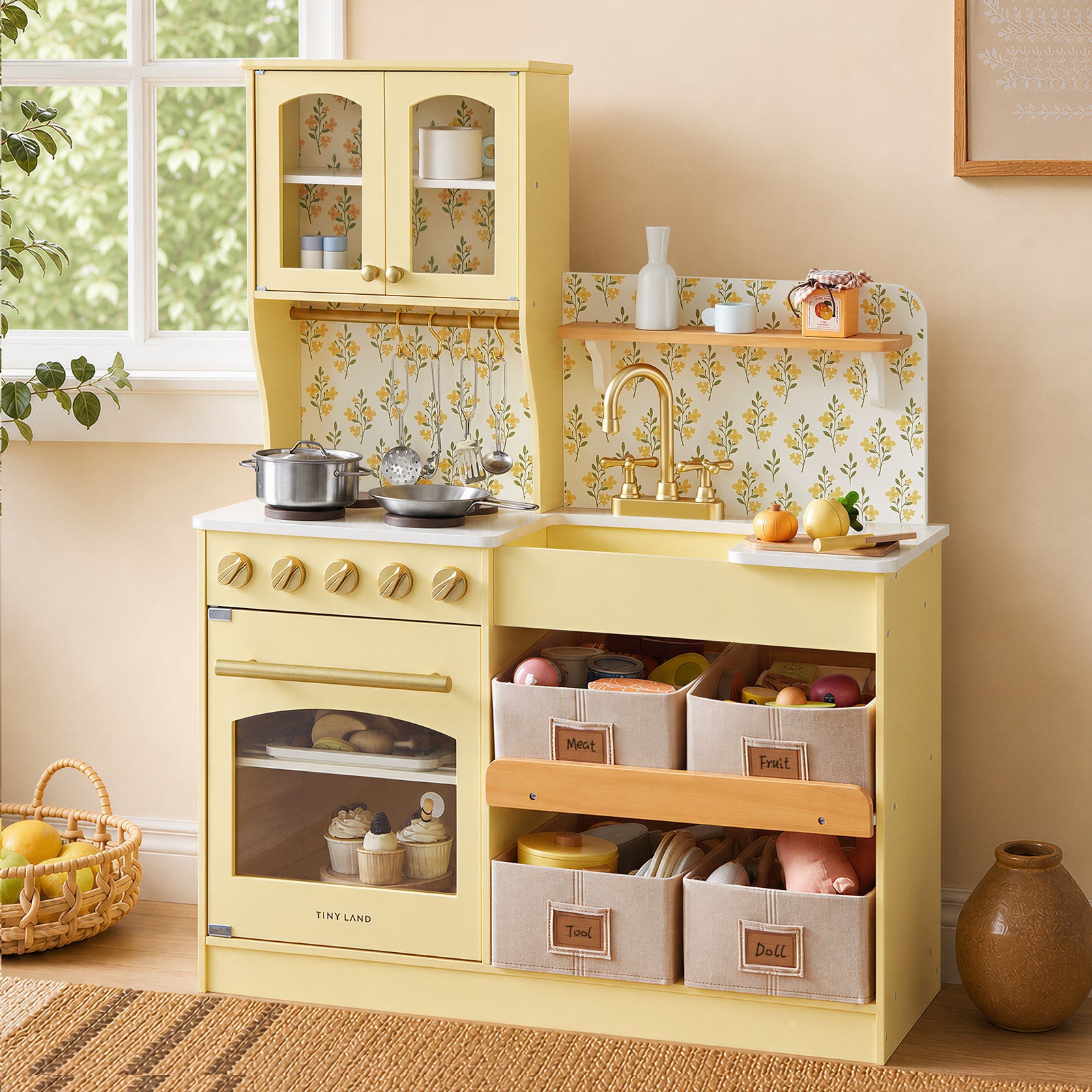 A children's wooden play kitchen in pale yellow, styled in a warm, retro-inspired room. The kitchen features a retro-style oven, a stovetop with gold knobs, a sink with a gold faucet, a floral yellow-and-white patterned backsplash, and three fabric storage bins labeled 'Fruit', 'Bakery', and 'Dishes'.