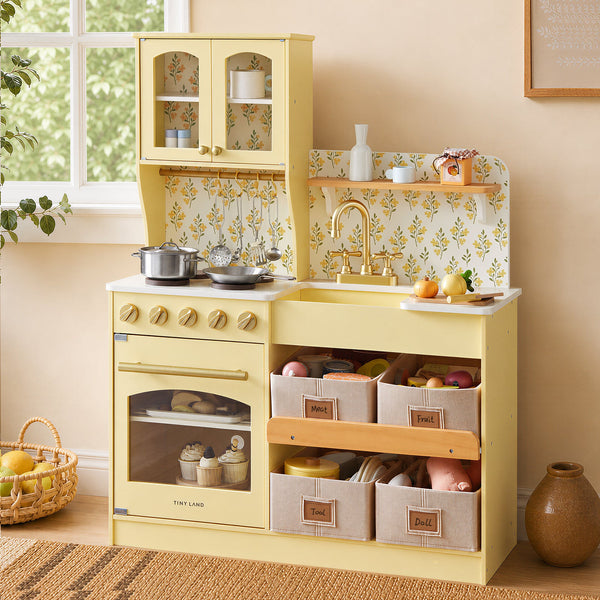 A children's wooden play kitchen in pale yellow, styled in a warm, retro-inspired room. The kitchen features a retro-style oven, a stovetop with gold knobs, a sink with a gold faucet, a floral yellow-and-white patterned backsplash, and three fabric storage bins labeled 'Fruit', 'Bakery', and 'Dishes'.