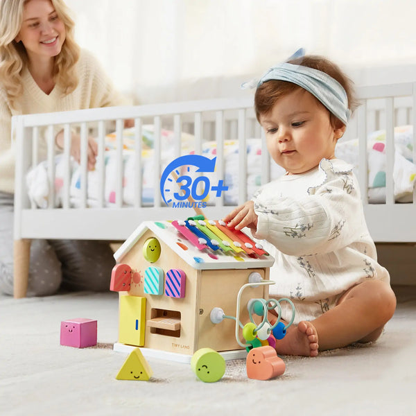 Baby playing with a colorful wooden toy in a nursery with a woman in the background.