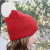 Person wearing a red knitted beanie with a white pom-pom against a blurred natural background