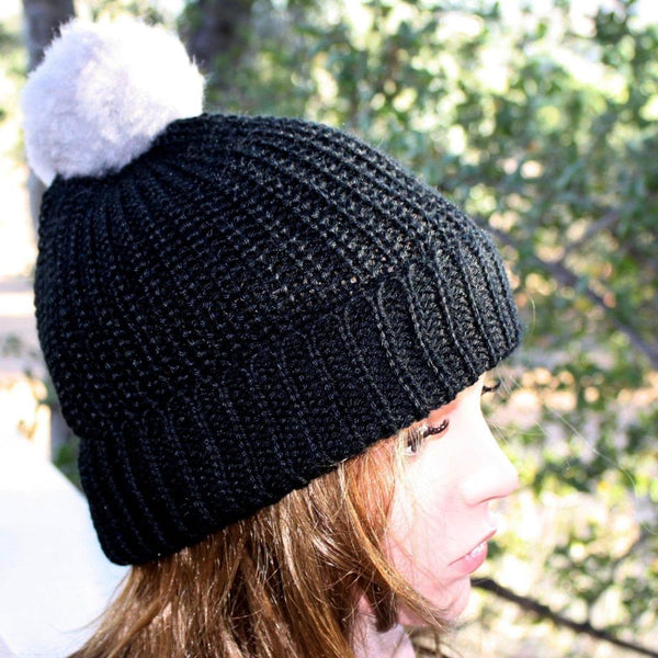 Black knit beanie with a white pom-pom worn by a person outdoors.