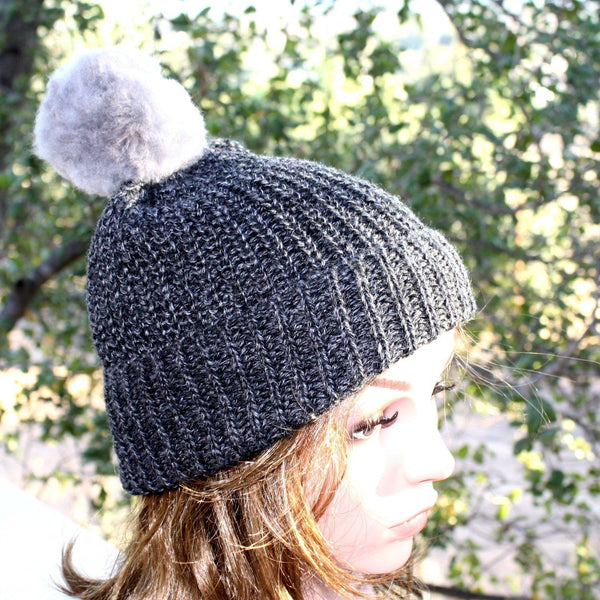 Person wearing a dark gray knit beanie with a pom-pom against a blurred natural background