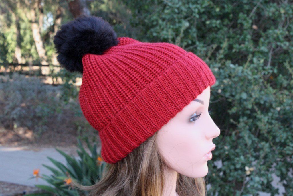 Red knitted beanie with a black pom-pom worn by a person outdoors.