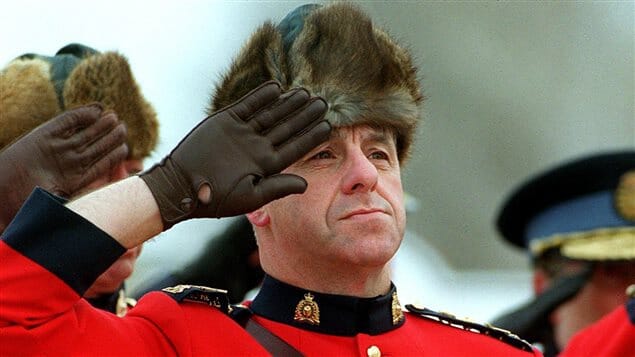 Person in a red uniform with a fur hat saluting, with another person in a similar uniform in the background.