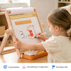 A young child in a cream dress is drawing on a wooden children's easel with a white board, marker, and small storage tray on a wooden table. The scene is brightly lit and set in a home-like environment.