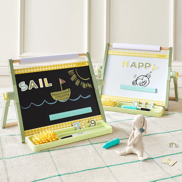 Two children's magnetic drawing easels displayed on a checkered rug in a bright room. The left easel has a black chalkboard panel with a yellow gingham border, showing the word 'SAIL' and a yellow boat drawing.