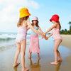 Three children holding hands on a beach with colorful hats and swimsuits.