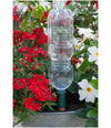 Clear plastic bottle with a green base used as a rain gauge in a garden with red and white flowers.