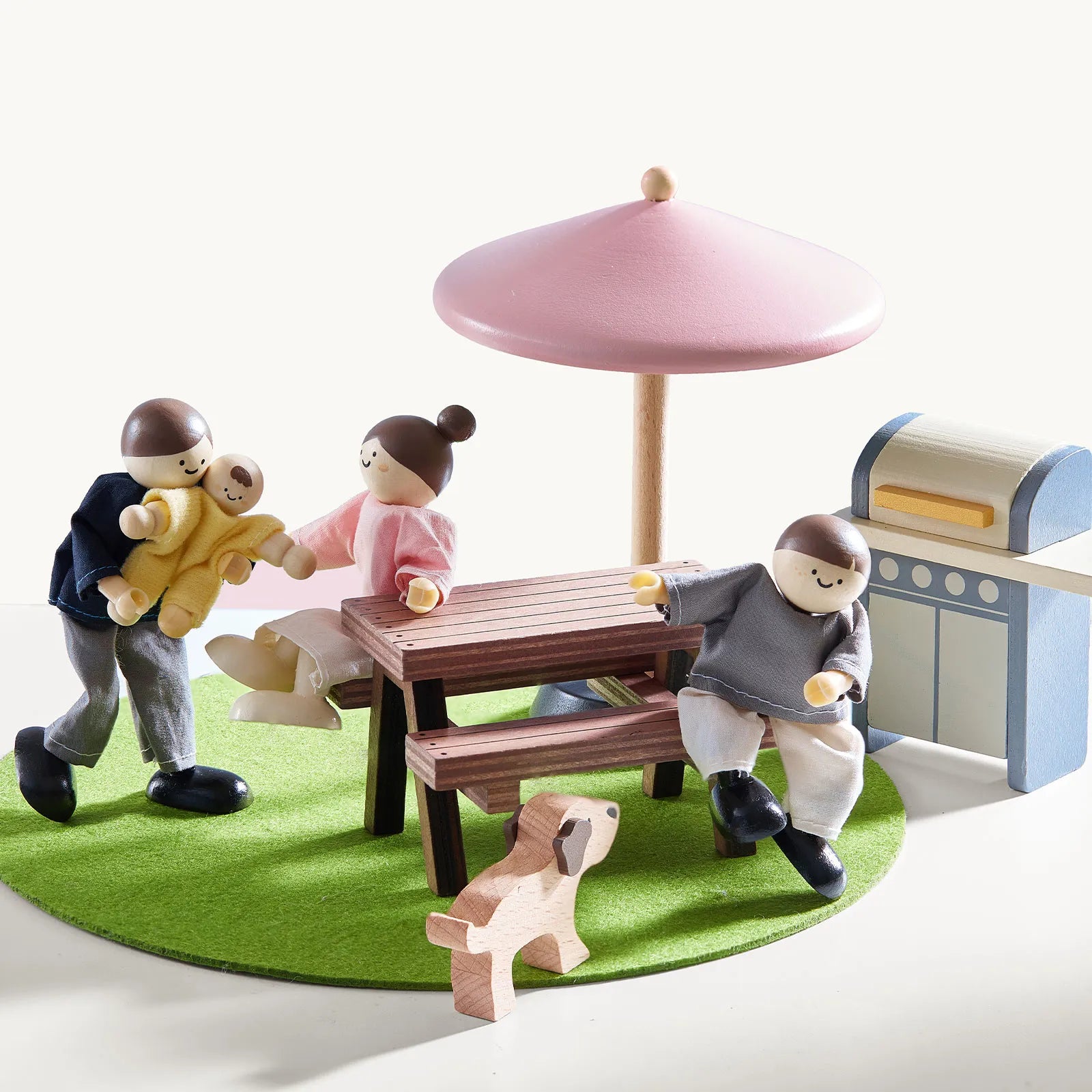 Toy figures of children and a dog around a table with a pink umbrella on a green mat.