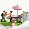 Toy figures of children and a dog around a table with a pink umbrella on a green mat.