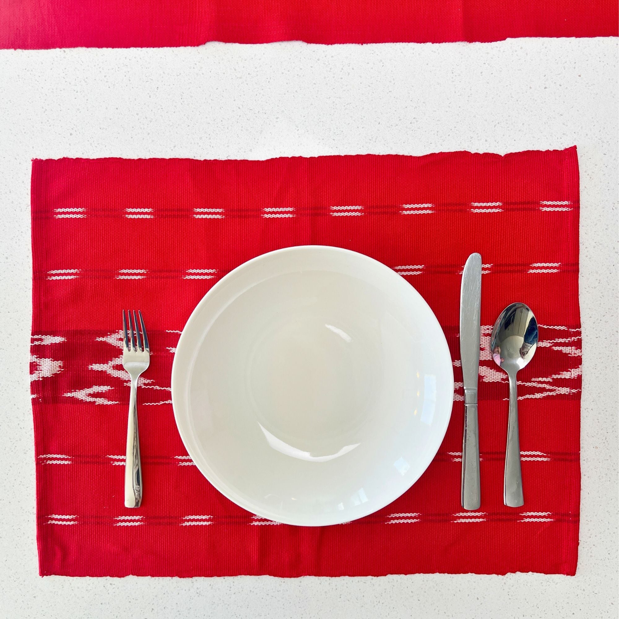 Red patterned placemat with a white plate, fork, knife, and spoon on a white background