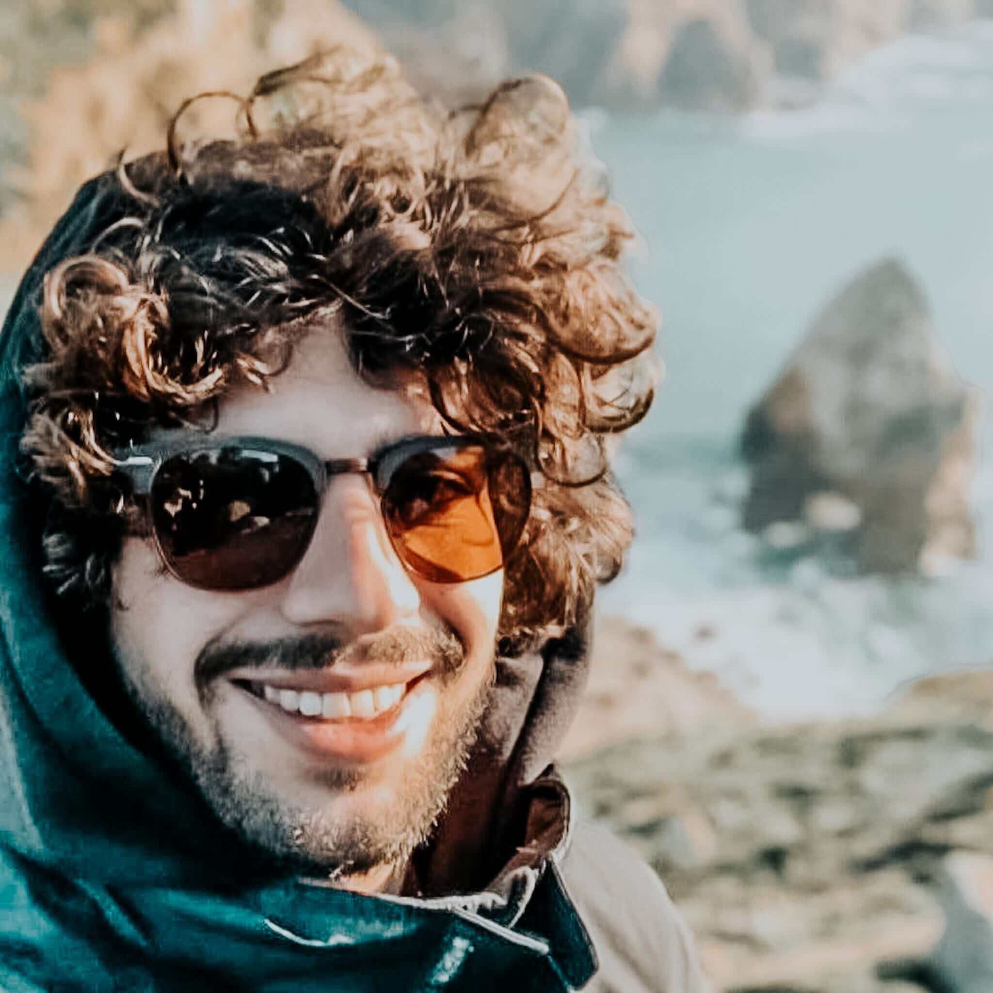 Man with sunglasses and curly hair standing by a body of water