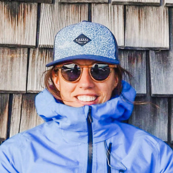 Person wearing a blue cap and jacket with sunglasses, standing against a wooden wall.
