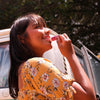 Woman applying lipstick in front of a car with trees in the background. Nourishing pitahaya & vitamin E lip balm with shea butter & coconut oil — repairs dry, chapped lips naturally. Plastic-free, handmade & chemical-free. Shop now.