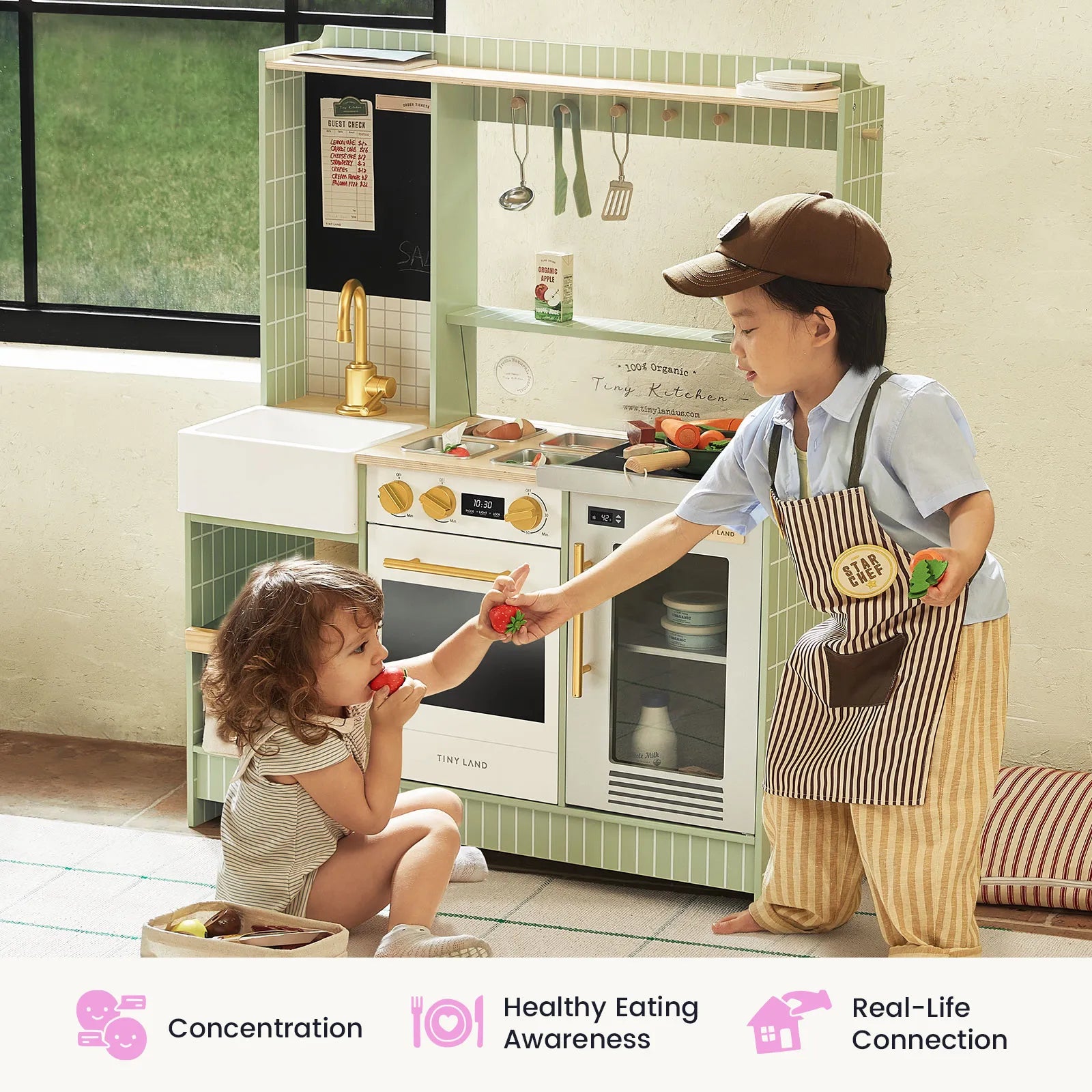 Two children playing with a toy kitchen set in a bright indoor room. One child stands wearing a brown apron and cap while feeding a toy strawberry to the other child, who is kneeling.