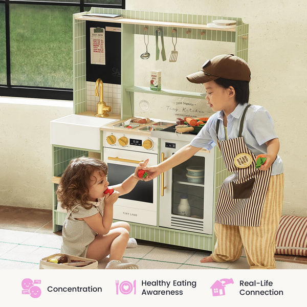 Two children playing with a toy kitchen set in a bright indoor room. One child stands wearing a brown apron and cap while feeding a toy strawberry to the other child, who is kneeling.