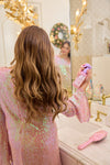 Woman in a sequin robe applying hair product in front of a bathroom mirror.