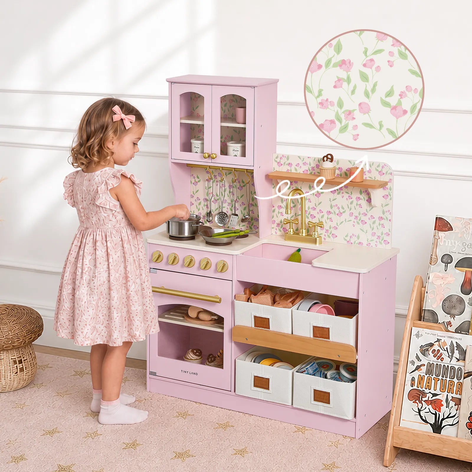 A young girl in a pink floral dress stands at a pink wooden play kitchen with floral backsplash, gold faucet, and storage drawers, in a bright nursery setting.