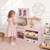 A young girl in a pink floral dress stands at a pink wooden play kitchen with floral backsplash, gold faucet, and storage drawers, in a bright nursery setting.