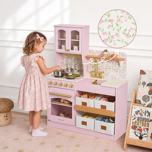 A young girl in a pink floral dress stands at a pink wooden play kitchen with floral backsplash, gold faucet, and storage drawers, in a bright nursery setting.