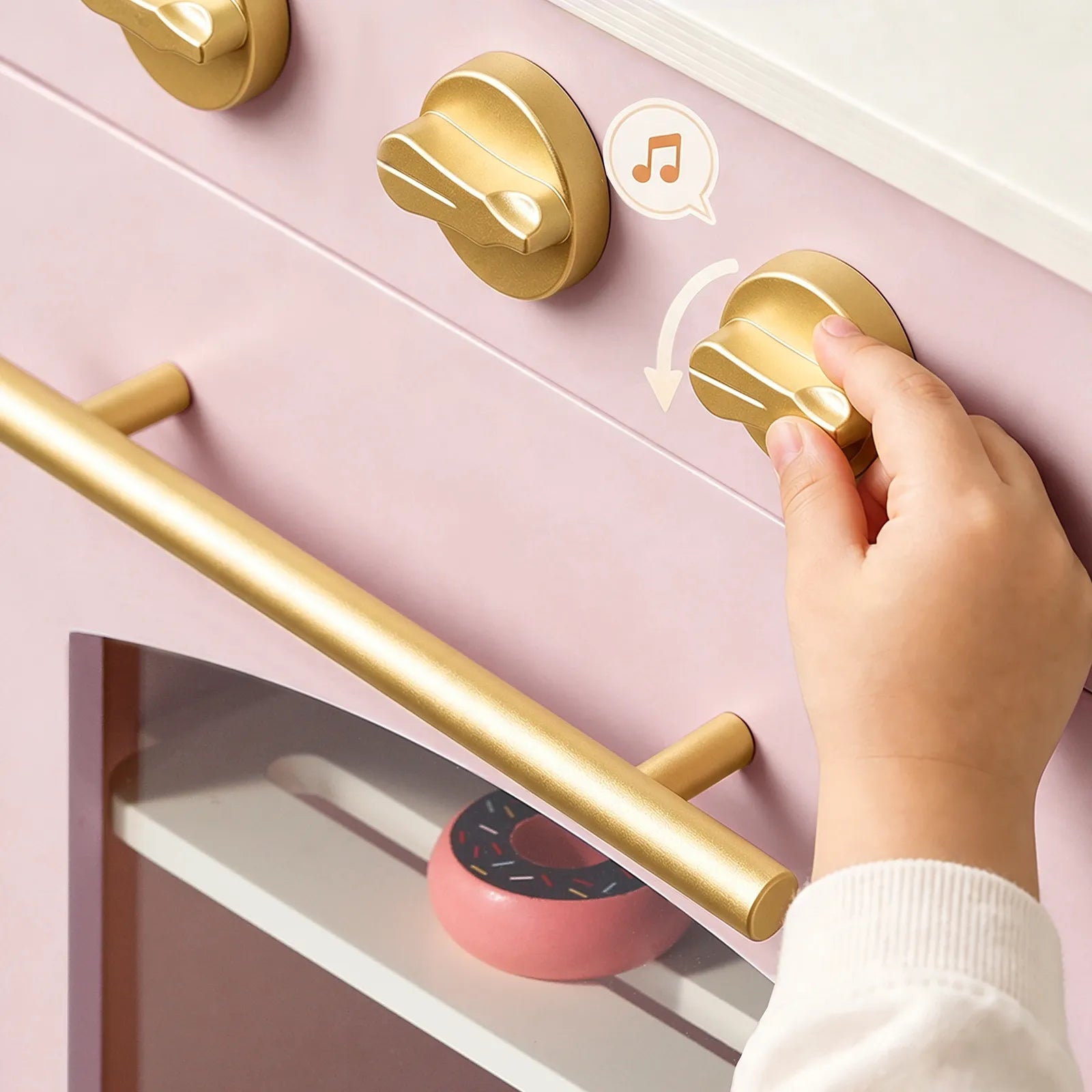 A child's hand turns a gold control knob on a pastel pink toy oven, with a small pink donut visible inside.