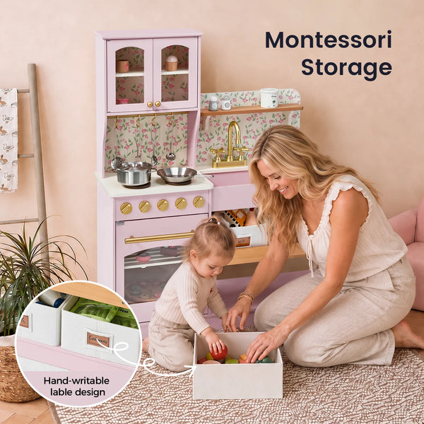 A woman and child are organizing toys in pastel pink Montessori storage boxes on a woven rug, with a matching pink play kitchen in the background. Text reads 'Montessori Storage' and a circular inset highlights the hand-writable label design.