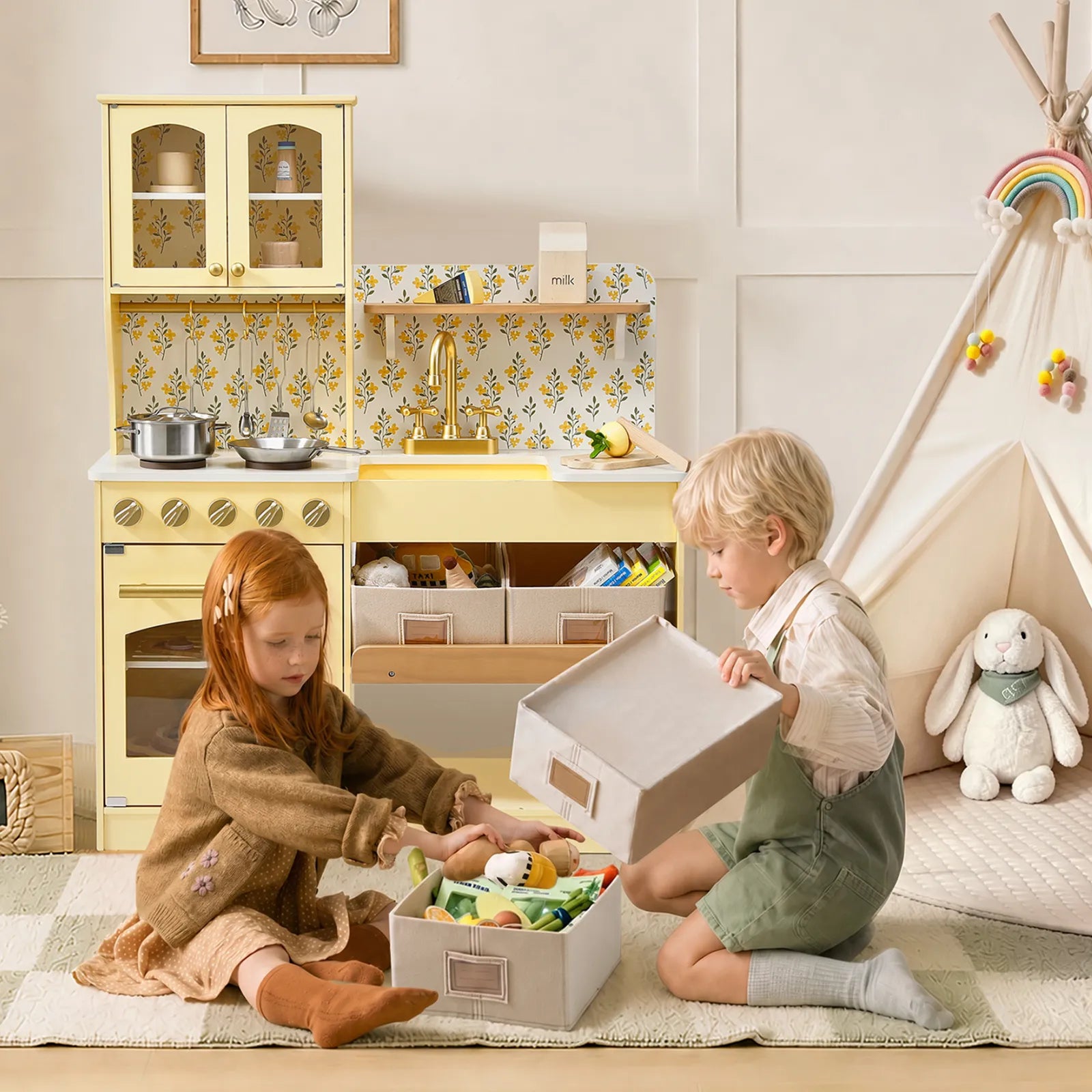 Two children playing with a pastel yellow wooden toy kitchen and storage boxes in a bright, styled children's room.