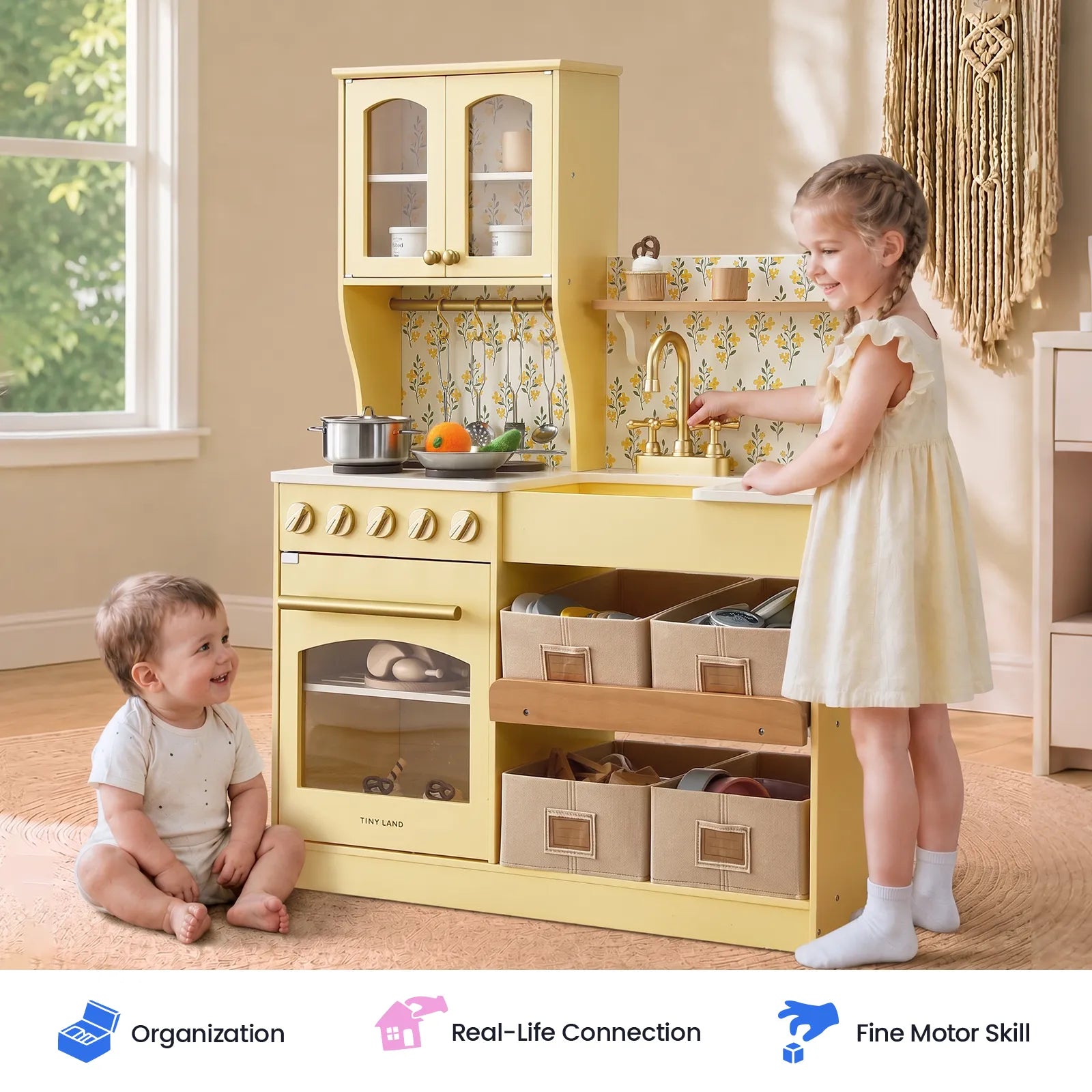 A children's wooden play kitchen set in a warm yellow color, styled in a bright room with two children interacting with it. One young girl stands at the sink, and a baby boy sits on the floor nearby.