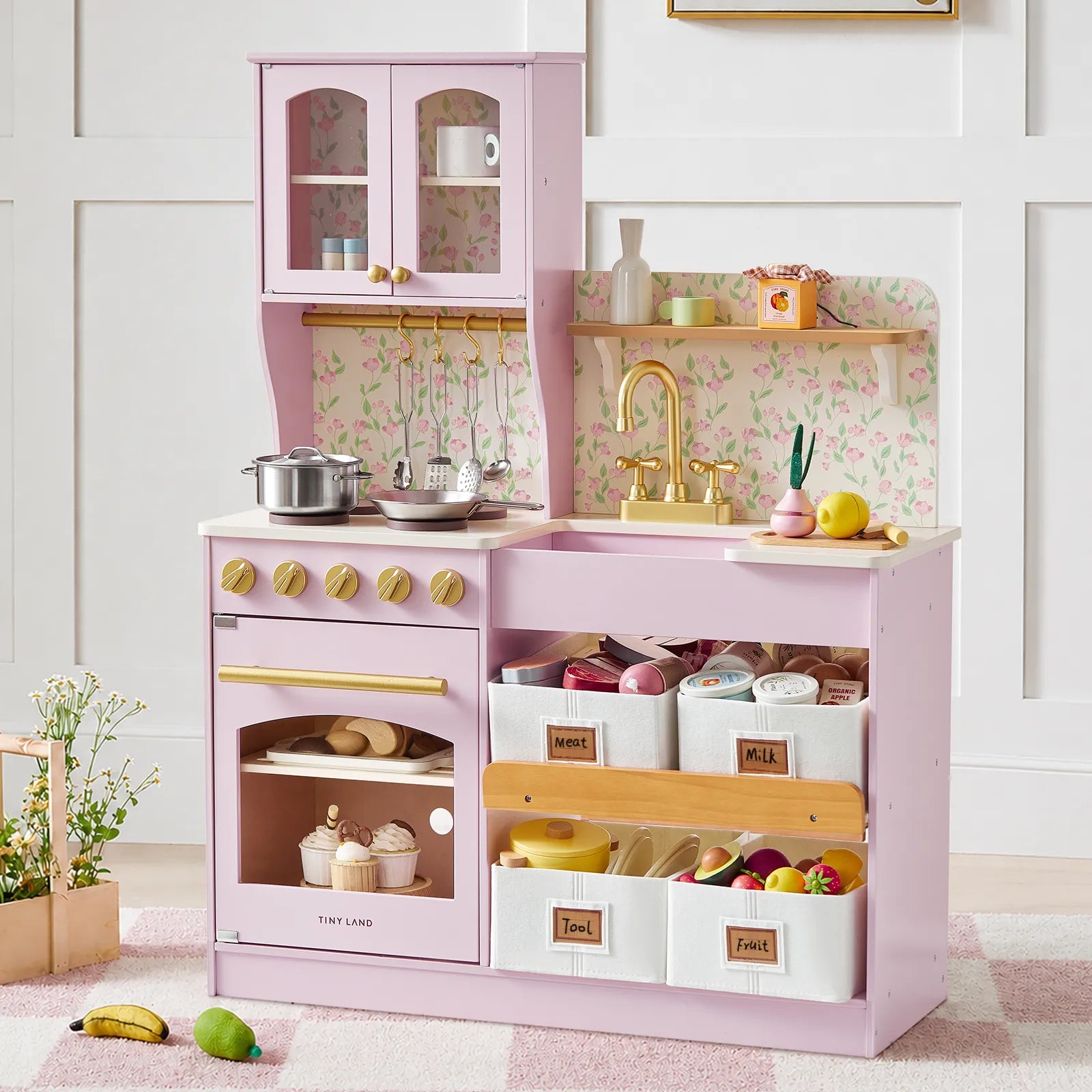 A pink children's play kitchen set with an oven, stove, sink, gold-tone faucet, floral backsplash, and storage bins filled with toy food and dishes, staged in a bright room with a rug and small plants.