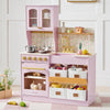 A pink children's play kitchen set with an oven, stove, sink, gold-tone faucet, floral backsplash, and storage bins filled with toy food and dishes, staged in a bright room with a rug and small plants.
