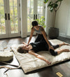 Two women in a massage or therapy session on a mat in a room with large windows and plants.