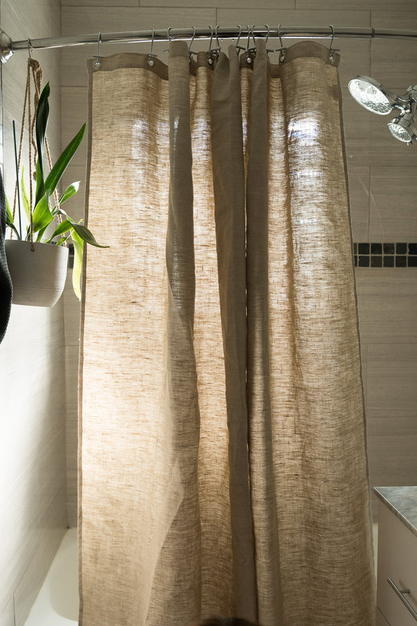 Beige textured shower curtain in a bathroom setting with a plant and light fixture.
